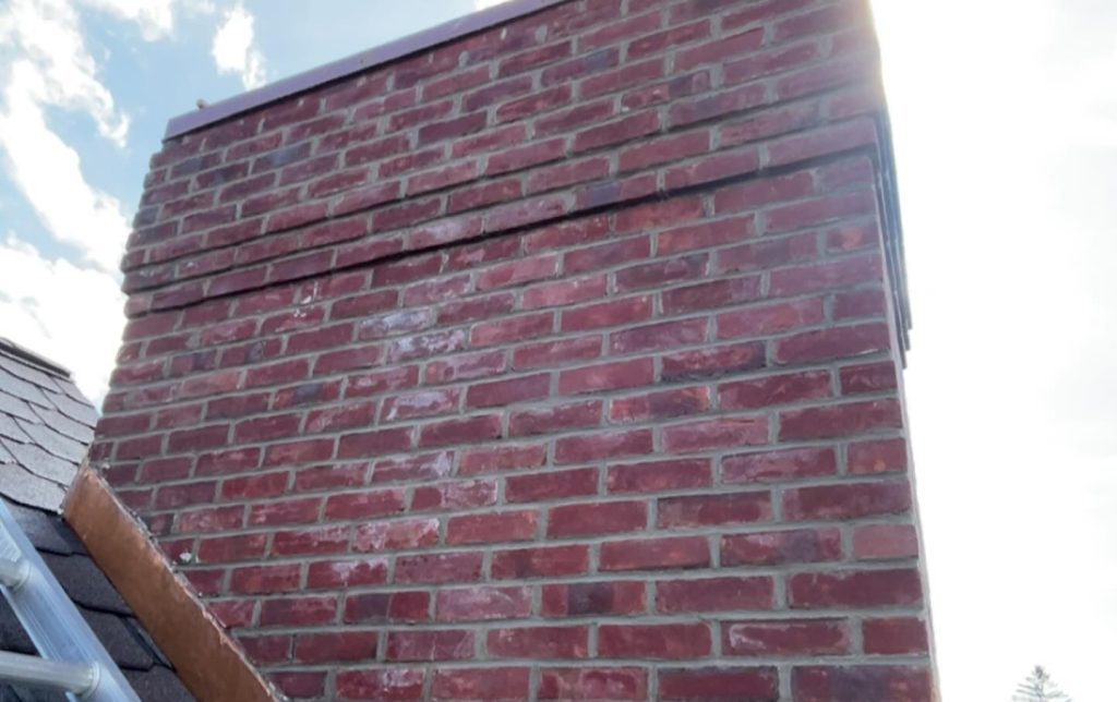 Red brick chimney with visible horizontal black line near the top, set against a partly cloudy sky. Sunlight creates a bright halo effect.