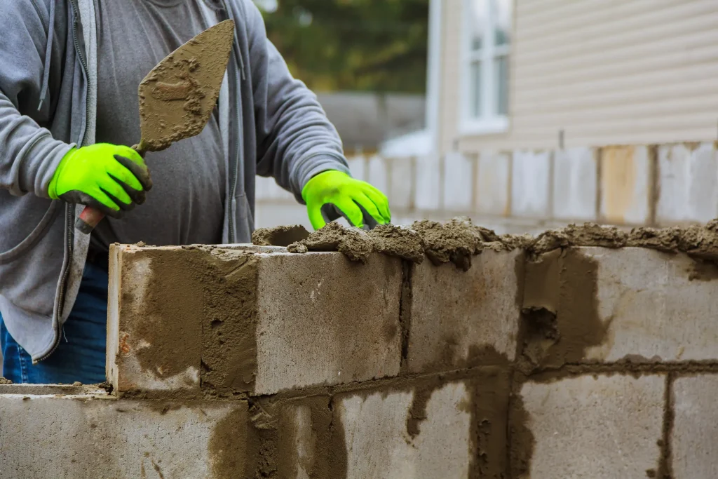 A construction worker wearing bright green gloves uses a trowel to apply mortar between concrete blocks, building a wall. The scene conveys focus and precision.