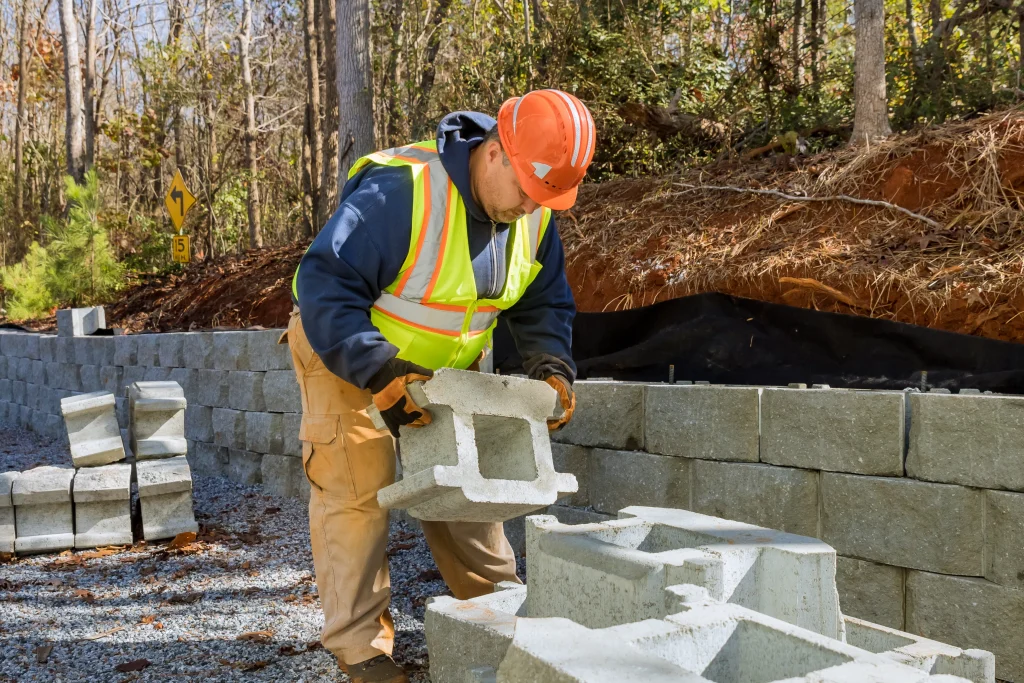 A construction worker in a hard hat and safety vest carefully places concrete blocks, building a retaining wall beside a forested area. He appears focused.