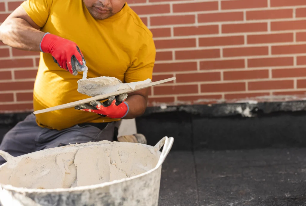 Man in a yellow shirt and red gloves spreads mortar on a trowel. He's near a brick wall, with a bucket of mortar on the ground, focused on his task.